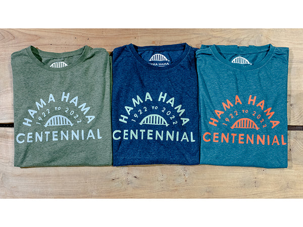 Three t-shirts are folded in half on a wood grain background. All three have the text "Hama Hama 1922 to 2022 Centennial" around a picture of the iconic Hama Hama bridge logo. 