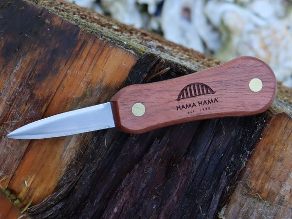 A oyster knife sits on a wooden log, the handle is rosewood with brass rivets and the blade is a hardened stainless steel. There are oyster shells in the background.,