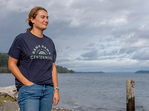 A person stands with their hair tied back and a hand in their pocket in front of the Hood Canal. They are wearing a lapis-blue t-shirt with a light green imprint that reads "Hama Hama 1922 to 2022 Centennial" around the iconic Hama Hama bridge logo. 
