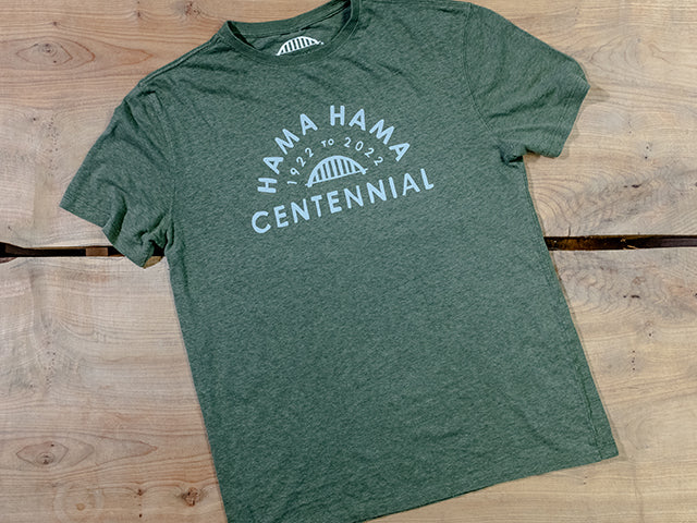 A cypress-green tee shirt lies on two wooden boards. The cool grey imprint reads "Hama Hama 1922 to 2022 Centennial" around a picture of the iconic Hama Hama bridge logo. 