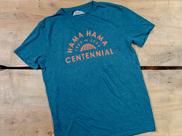 A teal tee shirt lies on two wooden boards. The salmon colored imprint reads "Hama Hama 1922 to 2022 Centennial" around a picture of the iconic Hama Hama bridge logo. 