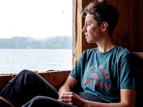 A person with short hair sits looking out a wooden framed window on to water and moutains. They are wearing a teal tee shirt with "Hama Hama 1922 - 2022 Centennial" written around the iconic Hama Hama bridge logo in a brilliant red/orange.   