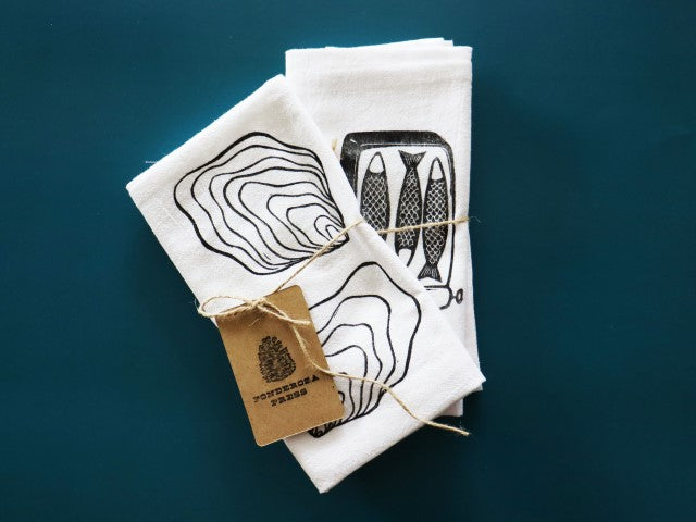 Two white cotton tea towels sit folded on a teal background. The one ion top has a block print in black ink of two oysters shells, the one in back has a block print in black ink of a tin with three small fish inside. They are tied with a hemp string and a tag that reads Ponderosa Press with the image of a pinecone above. 