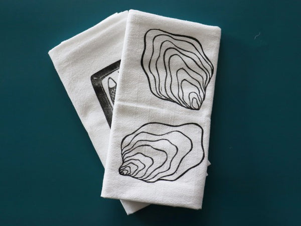 Two white cotton tea towels sit on a teal background. The one on top has block printed in black ink the image of two oyster shells, the one in back is partially obscured but has a block print with black ink of a open tin of fish.