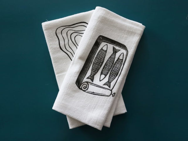Two cotton tea towels sit on a teal background. The one on top has a block print in black ink of a roll top tin can holding three small fish, the one in back is partially obscured but has a block print in black ink of two oyster shells. 